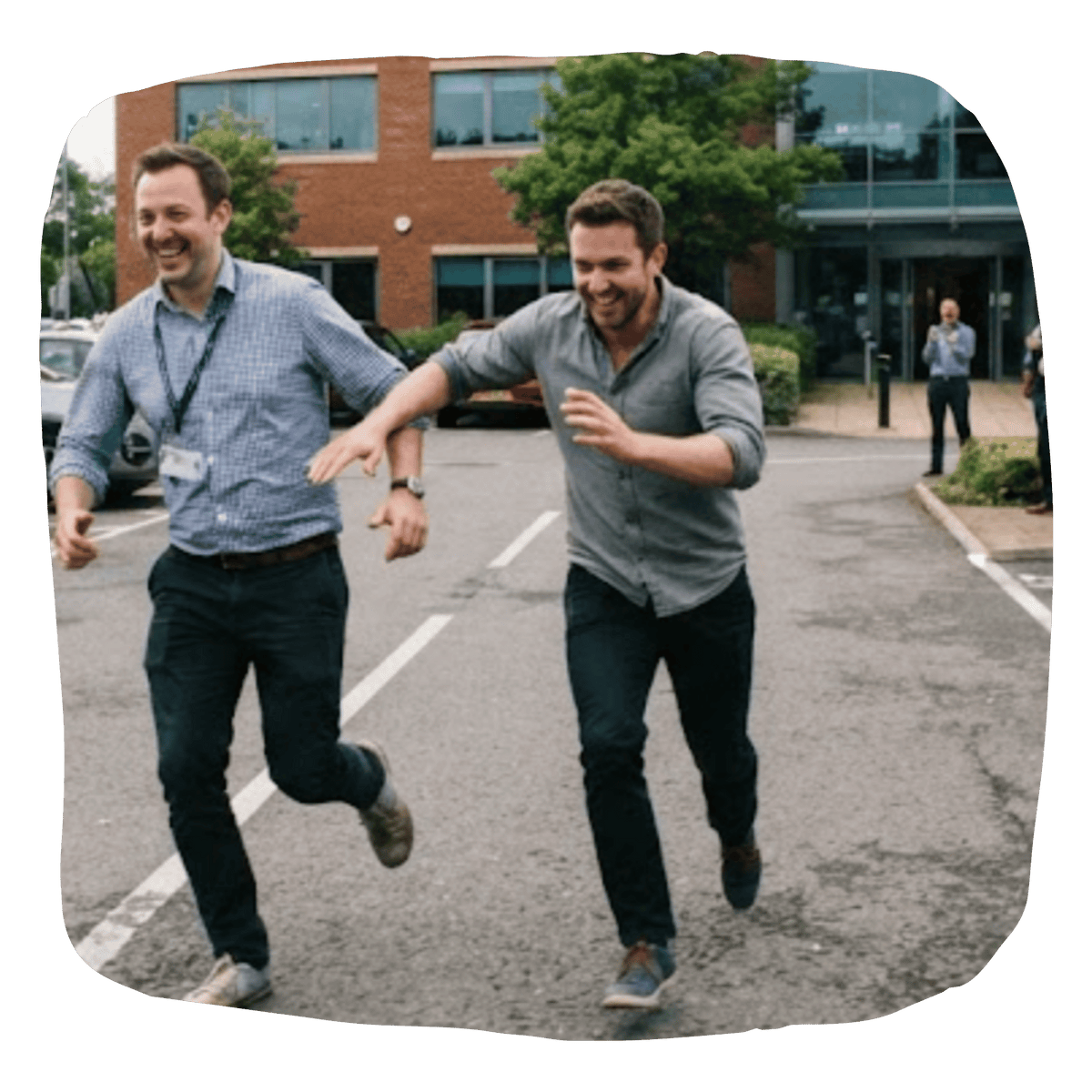 Two men running and laughing in an outdoor parking lot near a brick office building, with one wearing a light blue checkered shirt and the other wearing a gray button-up shirt.