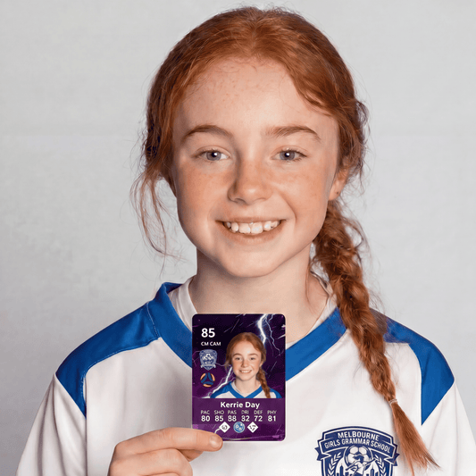 A smiling young girl with red hair in a braid holds up a purple player card featuring a pro squad prints design soccer card with her name and statistics. She is wearing a white and blue sports jersey against a light gray background.