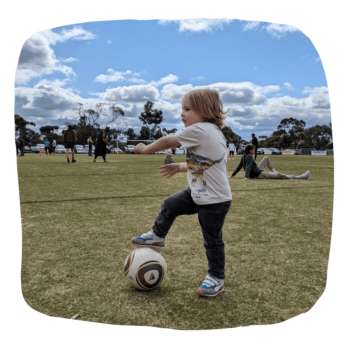 A young child wearing a white graphic t-shirt, dark jeans, and sneakers balances on a soccer ball on a grassy park field under a blue sky with clouds.