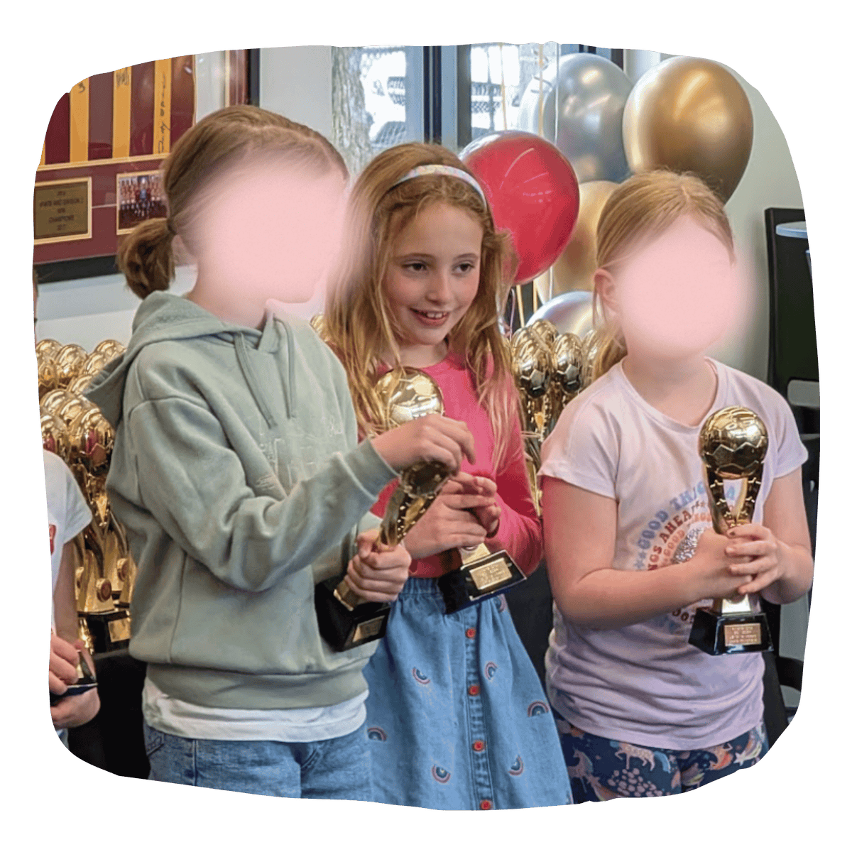 Three children standing together and holding shiny gold trophies in an indoor celebration setting with balloons in the background. The faces are blurred, and no visible brand logos are present.